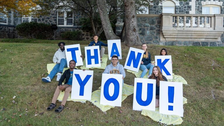 CSS Students pose in front of Tower Hall with the Give to CSS Day Towels and a sign that says thank you.