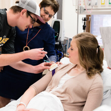 Male student practicing giving shots in St. Scholastica's Nursing Simulation Lab while the instructor and other students observe.