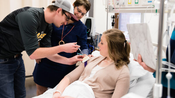 Male student practicing giving shots in St. Scholastica's Nursing Simulation Lab while the instructor and other students observe.